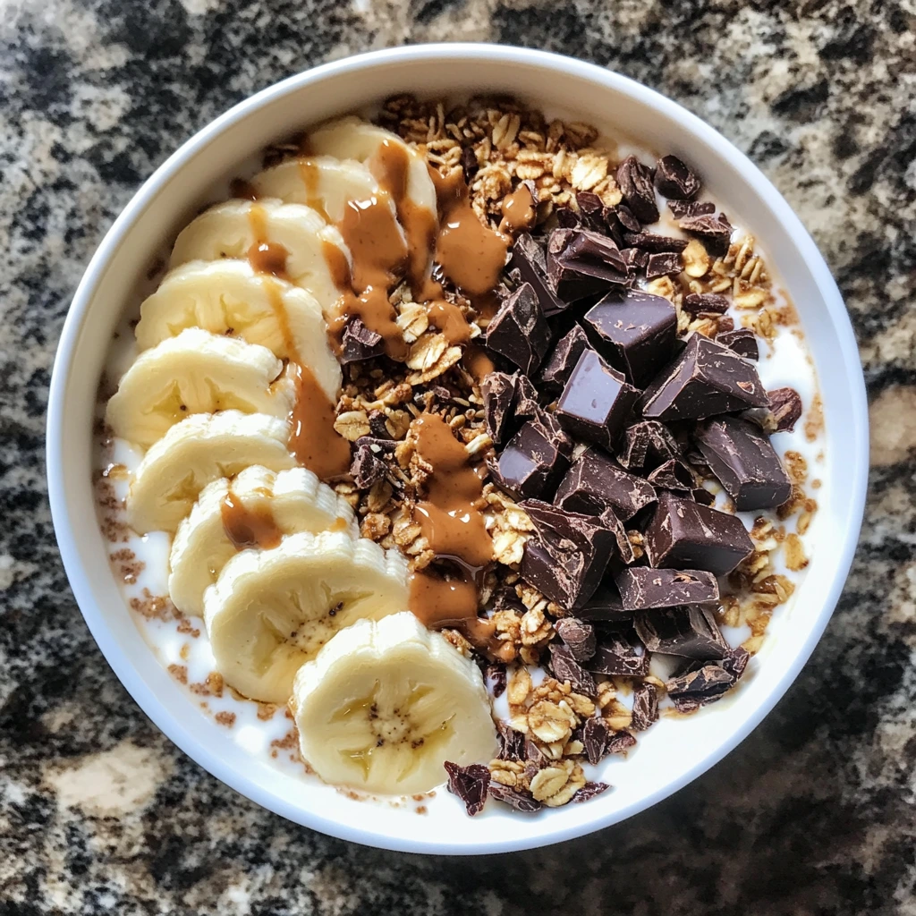 A rich and creamy Greek yogurt bowl topped with cacao nibs, dark chocolate chunks, sliced bananas, granola, and a drizzle of peanut butter.